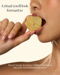 Close-up of a woman's hand and lower face holding a slice of citrus fruit near her mouth, with text reading 'A ritual you'll look forward to' and 'Blood Orange & Lemon Verbena Flavoured' on a warm cream background.