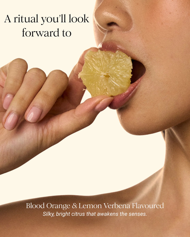 Close-up of a woman's hand and lower face holding a slice of citrus fruit near her mouth, with text reading 'A ritual you'll look forward to' and 'Blood Orange & Lemon Verbena Flavoured' on a warm cream background.